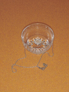 A transparent glass bowl sits on an orange surface, casting subtle reflections. It features intricate etched patterns along its sides. Draped gracefully over the bowl’s edge is a delicate silver chain, creating loops and gentle curves on the surface below. The composition highlights a contrast between the solid glass and the fluidity of the chain’s lines.