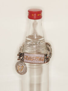 A clear bottle with a red cap is topped with a heavy chain featuring a detailed, gem-encrusted pendant and buckle. The design on these metallic accessories is intricate, featuring small, closely-set gems that add a sense of luxury and craftsmanship. The shiny surface of the chain and pendant contrasts with the bottle's smooth, transparent appearance.