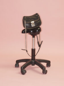 A sleek black swivel chair featuring a polished metal stand and caster wheels holds a stylish black handbag adorned with silver studs and chains. The handbag is structured with a metallic clasp closure. Set against a soft pink background, the composition blends industrial and fashion elements, creating a striking and chic visual contrast.