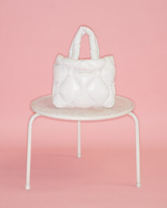 A white, quilted handbag with a puffy texture is placed on a small, round, white metal table with mesh details. The background is a solid, soft pink, creating a contrasting backdrop that highlights the bag. Two handles extend upward from the bag, which has a modern, stylish appearance, combining elegance with a casual, chic vibe.