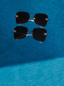 Two pairs of sleek, square sunglasses with dark lenses rest on a vibrant blue surface. The frames have a metallic detail on the sides, adding a touch of elegance. Light creates a soft diagonal shadow across the scene, enhancing the contrast between the glossy lenses and the textured background. The arrangement is symmetrical and visually striking.