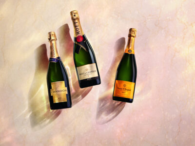 Three bottles of sparkling wine lie on a marble surface. Each bottle is elegantly designed, with gold foil wrapping the necks, accentuating their premium quality. The labels exhibit rich hues of deep green, black, and orange. Soft light casts gentle shadows, enhancing the luxurious ambiance and highlighting the subtle textures of their glass and the marble.