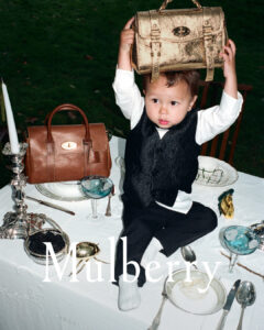 A child in formal attire stands on a lavishly set table outdoors, lifting a shiny gold handbag overhead. The table is adorned with fine china, decorative silverware, and crystal glasses. Beside the child sits a brown handbag. The background is a lush green lawn, adding contrast to the elegant yet playful scene.