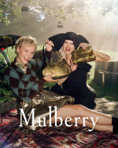 Two smiling individuals sit outdoors on a patterned rug, each holding a golden handbag. One wears a large black hat, while the other sports curly blonde hair and a patterned sweater. Sunlight filters through the leaves of a tree above, creating a warm and inviting atmosphere. The setting appears casual and relaxed, with subtle hints of nature's beauty.