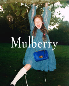 A cheerful person joyfully hangs from a tree branch, surrounded by lush green foliage. They're dressed in a flowing blue dress with sheer sleeves and white knee-high socks. The individual carries a vibrant blue bag with a distinctive gold clasp. The scene captures a playful, carefree moment set against a natural, tranquil backdrop.