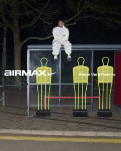 A person dressed in a white outfit sits relaxed atop a bus stop shelter, surrounded by leafless trees under the night sky. Highlighting the scene are three yellow training dummies standing below, contrasting against the darker background. The individual's footwear, bright and sporty, draws attention against the muted tones of the shelter and surrounding area.