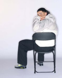 A person sits on a black folding chair, facing slightly backward. Wearing a light gray hooded jacket and black pants, they rest their arms on the chair's backrest. Their sneakers have a gray base with a neon green accent. The person's gaze is directed to the side, their hair styled in a sleek manner, against a neutral backdrop.