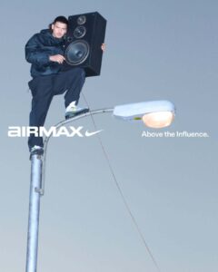 A person dressed in a dark outfit is perched on a high street lamp, holding a large speaker. The lighting is dim, giving a dusk-like appearance. The shoes have a distinctive design with white and green accents. A thin cable extends downwards, contrasting against the soft gray background, creating an urban and adventurous atmosphere.
