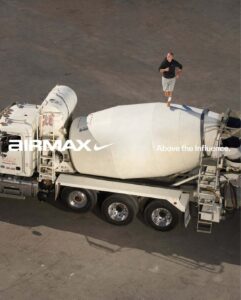 A man is running on top of a large cement mixer truck, balancing carefully on the rotating drum. The vehicle is parked on a wide, open expanse of concrete. Sunlight casts shadows on the ground, highlighting the shiny surface of the truck's metal components and the runner's athletic stance, with his shadow also visible beside him.