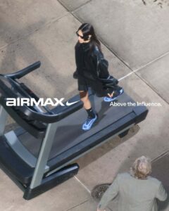 A woman in black sunglasses, a dark oversized jacket, and sport shoes walks confidently on a treadmill positioned outdoors. The scene captures her in motion, with her hair flowing and arms slightly extended. Her blue shoes stand out against the muted colors of the concrete sidewalk. A person in a gray jacket is visible nearby, glancing upward.