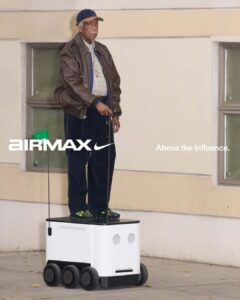 An elderly man stands on a small, box-like platform with wheels, resembling an autonomous delivery robot. He wears a leather jacket, a cap, and holds a cane. The platform features facial-like circular designs. The setting is an outdoor urban area with beige walls and windows, creating a contrast between the modern device and traditional attire.