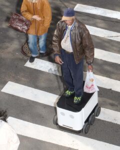 A man stands atop a wheeled robotic platform, holding a white bag in one hand and a control device in the other. He wears a brown leather jacket, a cap, and neon green shoes. Beside him, an individual in a tan jacket with a large handbag crosses a street, marked with wide white lines.