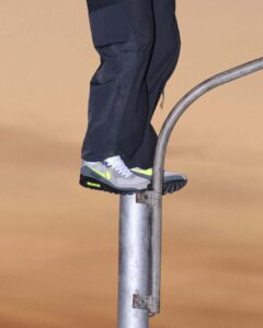 A person wearing dark pants and gray sneakers with neon accents is balancing on a metallic pole. The pole has a curved arm, suggesting a utility structure, set against a flat, beige background. The shoes feature a black sole, white laces, and the background's color hints at sunset or artificial lighting.