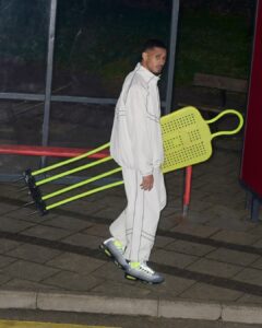 A person dressed in a light-colored tracksuit walks along a paved surface, holding a large, perforated, bright yellow object with several extended spikes. The individual wears grey sneakers with neon green accents. There are reflective glass panes and a wooden bench in the background, along with a curved red metal rail nearby.