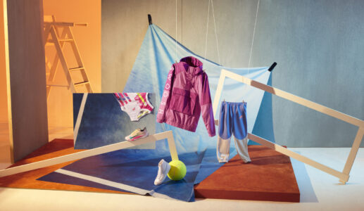 A vibrant composition showcases a purple jacket and blue pants displayed against a backdrop of suspended geometric shapes. A tennis ball and two shoes complement the scene’s dynamic arrangement. The background features bold blue and orange hues, with a ladder hinting at an artistic setup. The overall aesthetic combines elements of style and sport in a playful manner.