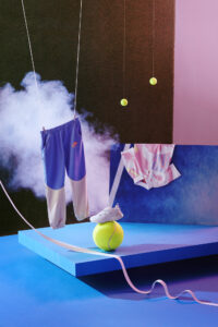 A surreal scene features a pair of shorts suspended by wires, accompanied by a shirt placed nearby. A sneaker rests atop a large, bright tennis ball, with a ribbon cascading down. Vibrant blue and pink surfaces dominate the background, while tennis balls and mist contribute to the dynamic, whimsical atmosphere.
