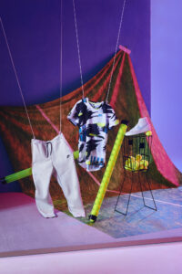 Artistic display with a colorful abstract background features suspended clothing and props. A pair of light-colored pants and a multicolored shirt are creatively hung, with a vibrant cylindrical object beside them. A mesh basket holds tennis balls beneath a displayed casual shoe. The setting's bold colors evoke a playful, dynamic atmosphere using modern, stylish elements.