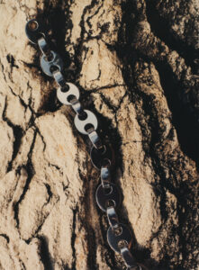 A textured background resembling rough, craggy rock features prominently, with a metallic chain draped across it. The chain consists of alternating oval and circular links, reflecting light in a polished, metallic sheen. The contrast between the smooth, shiny surface of the chain and the rugged, uneven surface underneath creates a striking visual juxtaposition. Shadows add depth and dimension.