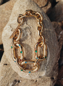 A gold chain bracelet rests elegantly atop a textured rock surface, glistening in soft, natural light. The links are embellished with small, colorful gemstones, including green, blue, and red. The intricate design and polished finish contrast beautifully against the rugged stone backdrop, showcasing the blend of opulent craftsmanship and earthy elements, creating a striking visual harmony.