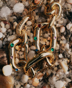 Golden chain links adorned with vibrant, colorful gemstones rest among smooth, wet pebbles. The stones range in color, including emerald green, deep blue, and rich purple. Bubbles cling to the metallic surface, enhancing its shine and creating a luxurious contrast against the muted, earthy tones of the surrounding rocks, suggesting a lavish, natural setting.