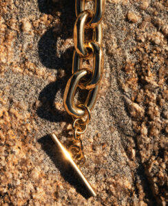 Golden, chunky chain links glint under direct sunlight, casting soft shadows on a textured, sandy surface peppered with small, rough stones. The polished metals form a T-bar toggle clasp, reflecting light brilliantly. This luxurious, gleaming piece stands out against the natural, earthy background in rich gold and beige hues, exuding elegance and sophistication amidst the rugged terrain.