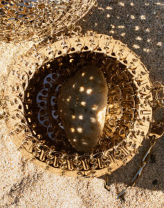 A rocky shoreline serves as a backdrop for an intricate metallic mesh structure. The mesh, circular and reflective, is adorned with linked components, encasing a smooth pebble at its center. Sunlight filters through, creating a patterned play of light and shadow. Surrounding grains of sand add texture, contrasting with the intricate metallic construction's ornate details.