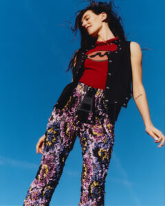 A person stands against a deep blue sky. They wear a red top under a black vest accented with jewels, alongside purple and yellow textured pants. A bold red necklace adorns their neck. Their hair flows freely with the breeze, and they pose with relaxed grace, evoking a sense of freedom and confidence.