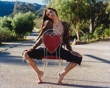 A person sits on a heart-shaped chair in an outdoor setting, surrounded by trees and a clear sky. The individual has long hair and is wearing dark pants and a brown top. Their pose is relaxed and confident, with bare feet resting on the ground. The background features a sunny path and distant hills.