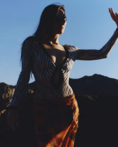 A person is framed against a deep blue sky, standing in a silhouetted pose. The backdrop features a rugged, mountainous terrain. The individual wears a striped top with a deep neckline and patterned orange wrap. Their arm is raised, partially shadowed by the bright sunlight, emphasizing the contours and features beautifully against the landscape.