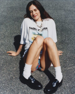 A young woman with long hair is seated on a sunlit pavement, eyes closed in a relaxed posture. She wears a white shirt featuring a graphic design, along with a necklace, black shoes, and white socks. Her legs are extended forward, and she appears content, enjoying a warm, tranquil moment outdoors. Shadows suggest a bright, clear day.