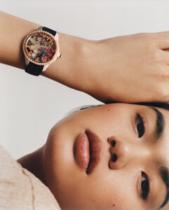 A woman lies on her side, resting her head on her hand. Her wrist displays an ornate watch with a colorful, intricate design and a black strap. Her expression is calm and neutral, with natural makeup accentuating her features. The focus on her watch and face suggests elegance and serene confidence amidst a simple background.
