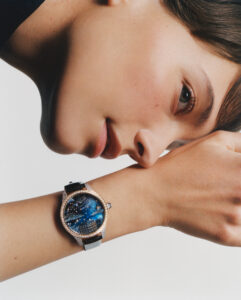 A person gazes thoughtfully, resting their head on an outstretched arm. Their skin is smooth and hair neatly styled. A luxurious wristwatch, featuring a celestial-themed design with a dark blue dial and starry patterns, adorns the wrist. The watch is encased in a shiny frame and secured by a sleek black strap, conveying elegance and sophistication.