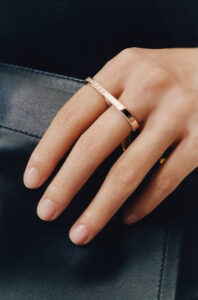 A close-up view captures a hand resting on a smooth, dark surface. The fingers are adorned with two sleek, elegant rings: one plain and the other subtly embellished with small details. The skin tone is a soft, natural hue, and the light highlights the gentle curves and texture of the hand and jewelry, evoking a sense of refinement and elegance.