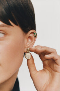 A person with short, dark hair is adorned with two earrings. One is an ornate, circular design held by their hand, featuring clear details. The other earring is smaller, located higher on the ear, with a colorful geometric pattern. The hand gently holds the larger earring, showcasing a neutral manicure. The background is softly blurred.