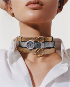 A person wears a white collared shirt with multiple layered metallic chokers. Each choker is uniquely detailed; some shimmering in gold and others in silver, featuring circular embellishments with textured designs. The overall look is elegant and stylish, highlighting a blend of modern fashion trends and classical accessory style, creating a striking visual appeal.