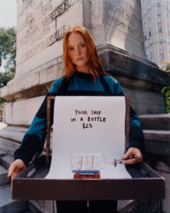A person with long red hair stands outdoors, holding a tray featuring a paper and a ship in a bottle. They wear a dark striped top. The background includes a historical stone monument with engraved text and a glimpse of urban buildings on a sunny day. Shadows and greenery suggest a peaceful, relaxed setting.