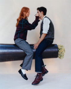Two individuals are sitting on a large cylindrical object adorned with flowers at one end. One, with long red hair, is applying makeup to the other, who wears a gray vest over a white shirt and dark jeans. The atmosphere is casual, with both wearing stylish yet relaxed attire, including checkered pants and maroon boots.