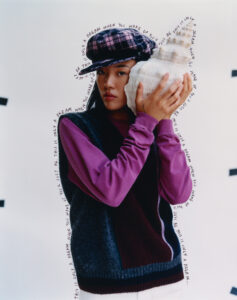 A person in a purple long-sleeve shirt wears a dark, patterned cap. They hold a large, spiral seashell to their ear. The background is light, and there are visual elements that resemble handwritten text arcing around them. The mix of soft colors and textures gives a dreamy, introspective feel to the composition.