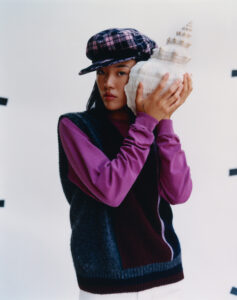 A person stands against a plain backdrop, holding a large seashell to their ear. They wear a checkered cap and a color-blocked sweater over a magenta top. The attire is casual yet stylish, with contrasting textures and patterns. The seashell, with its intricate spirals, adds an element of nature to the scene.