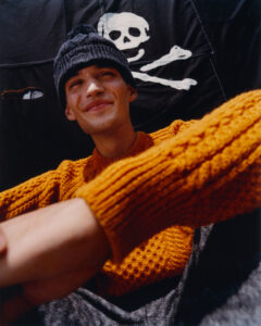 A person in a textured, orange sweater and dark knit hat sits comfortably, with an expression that hints at contentment or amusement. The background features a black fabric with a bold, contrasting white design of a skull and crossbones, adding an edgy or adventurous element to the scene. Shadows play across the figure’s face and clothing.