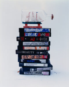 A collection of vintage VHS tapes, each with a uniquely designed spine, is stacked in an uneven pile. Atop this pile rests a decorative ship encased in a glass bottle, adding a nautical charm. The background is minimalistic, drawing focus to the colorful and eclectic assortment of visual media and the intricate model ship.