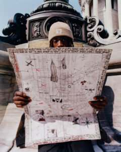 A person wearing a beige knit cap and dark clothing is holding an intricately drawn map with elements such as buildings and navigational markers. They are seated against an ornate architectural structure with elaborate scrollwork. The background reveals part of a grand building with columns and detailed stone carvings, suggesting an urban setting on a clear day.