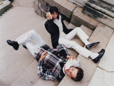 Two people are reclining on stone steps, creating a casual, fashionable tableau. One wears a dark coat with white pants and holds a large shell near their ear. The other, dressed in a plaid jacket, leans back playfully with a similar shell. Their expressions and relaxed poses contrast with the structured surroundings, blending whimsy with style.