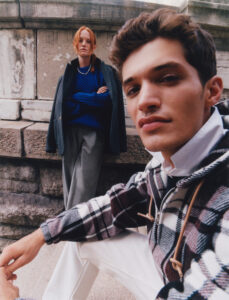 A young man and woman pose fashionably against a weathered stone wall. The man, wearing a plaid jacket and white pants, looks directly at the viewer with a confident expression. The woman, more distant, has red hair, wearing a blue sweater and gray coat. The setting evokes an urban, classic atmosphere with a touch of modern style.