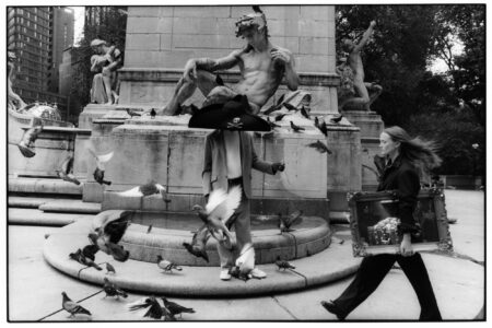 A person dressed in a suit stands near a fountain, surrounded by pigeons in flight. They wear a large hat, giving a whimsical touch to the scene. Another person walks past holding a framed picture, their hair captured mid-motion. Classical statues adorn the background, while trees and tall buildings frame the urban setting.