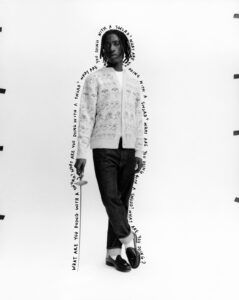 A person with medium-length hair stands confidently against a plain background. They wear a patterned cardigan, a white shirt, dark cuffed jeans, and loafers. Holding a sword casually by their side, their gaze is directed towards the viewer. Black tape pieces are visible around the edges, giving a sense of a staged set. Surrounding lines emphasize their presence.