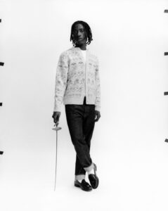 A person stands confidently, dressed in a patterned cardigan and rolled-up jeans, holding a slender sword. They wear loafers with socks, exuding a poised and stylish demeanor. The backdrop is plain, creating a minimalist and focused composition. Dark hair frames the face, contributing to the overall elegant and contemporary look, enhanced by the monochrome palette.