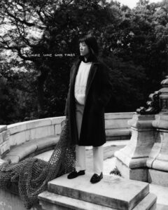 A person stands on a stone platform in a formal, long coat layered over a sweater and trousers. They appear contemplative, gazing into the distance. A textured cloth drapes over the edge of the structure. In the background, a lush canopy of trees creates a natural setting, blending with the classical architectural elements surrounding the figure.