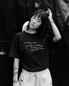A person stands in front of a dark backdrop, wearing a dark shirt and light-colored sweatpants. Their hand is in their hair, and they appear contemplative. Tattoos are visible on their arm, featuring abstract designs. The monochrome setting and soft lighting create a thoughtful and artistic atmosphere, highlighting the casual and relaxed pose of the individual.