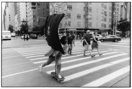 A person speeds across a crosswalk on a skateboard, holding a large flag with a skull and crossbones design. Pedestrians stroll nearby, seemingly unfazed. The scene captures urban energy, with a backdrop of towering buildings and cars on the street. The atmosphere is dynamic, blending everyday life with a touch of rebellion and adventure.